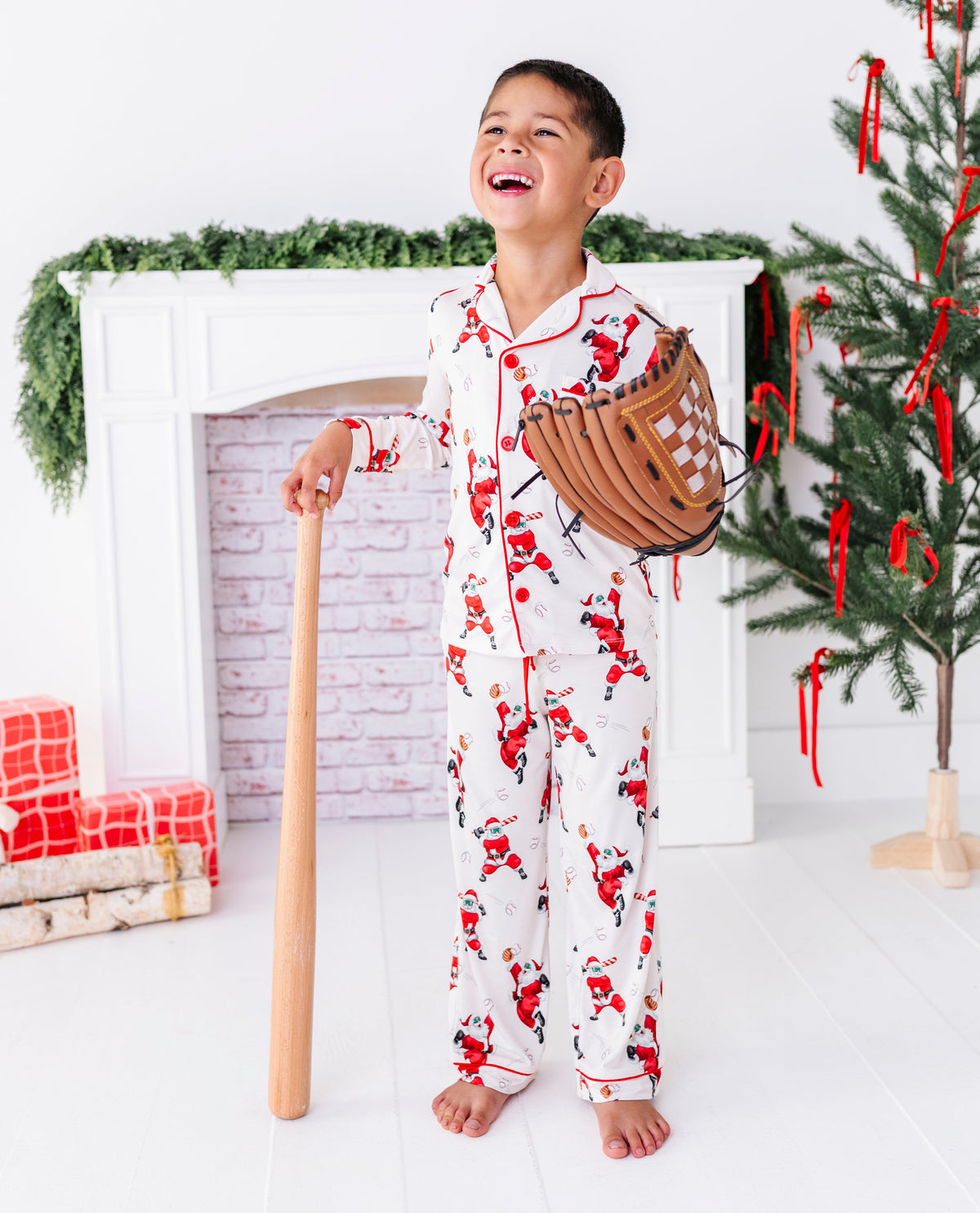 A little boy wearing a loose-fitting bamboo lounge set featuring Santa throwing baseballs and knocking them out of the park! He's holding a wooden bat and wearing a baseball glove while standing in front of a fireplace and a blurry Christmas tree