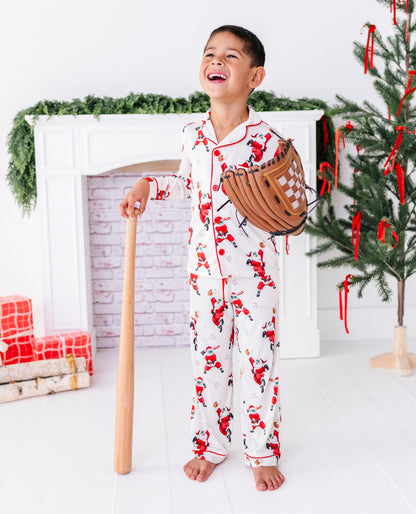A little boy wearing a loose-fitting bamboo lounge set featuring Santa throwing baseballs and knocking them out of the park! He's holding a wooden bat and wearing a baseball glove while standing in front of a fireplace and a blurry Christmas tree