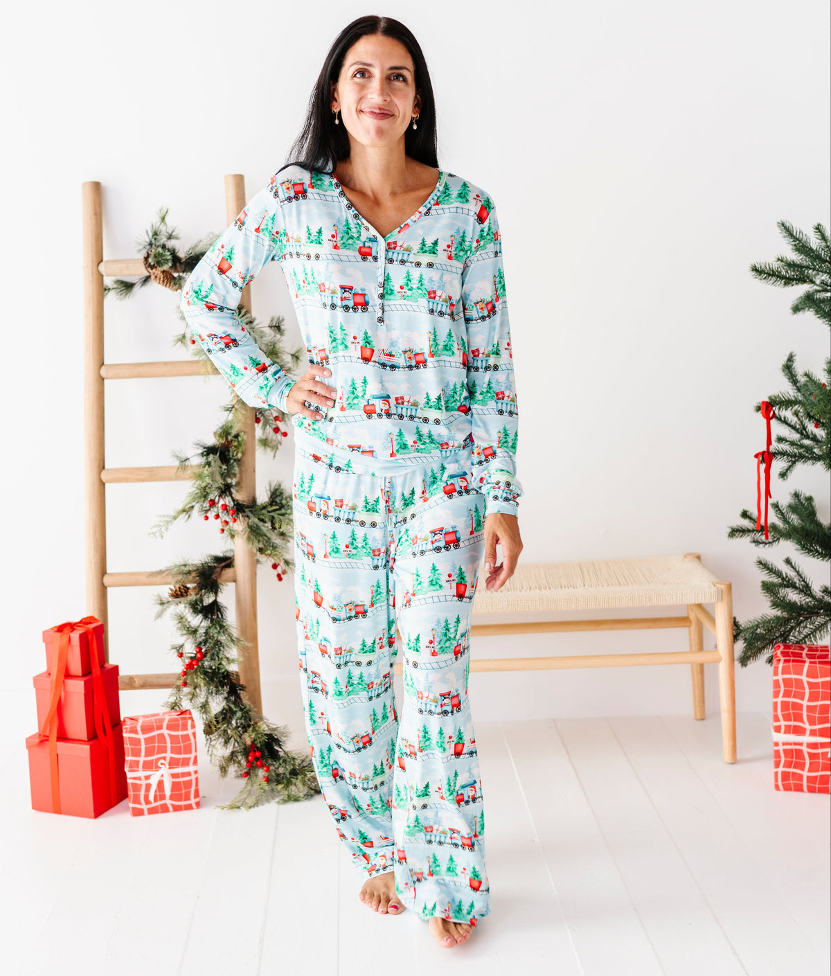 A woman stands in a bright white room wearing a matching long-sleeve pajama set with a Christmas tree and sleigh print. The pajamas have a light blue background with green trees, red and white sleighs, and small red ornaments.