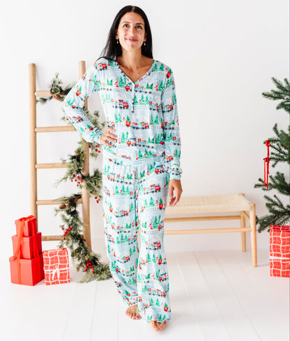 A woman stands in a bright white room wearing a matching long-sleeve pajama set with a Christmas tree and sleigh print. The pajamas have a light blue background with green trees, red and white sleighs, and small red ornaments.