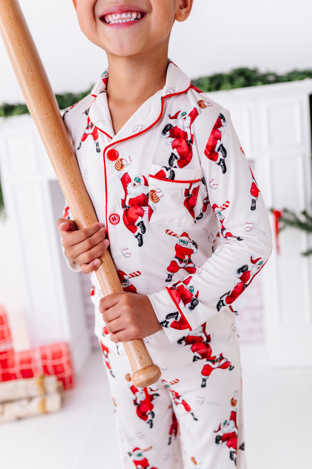 A close-up shot of a little boy wearing a loose-fitting bamboo lounge set featuring Santa throwing baseballs and knocking them out of the park! He's holding a wooden bat while standing in front of a fireplace and a blurry Christmas tree