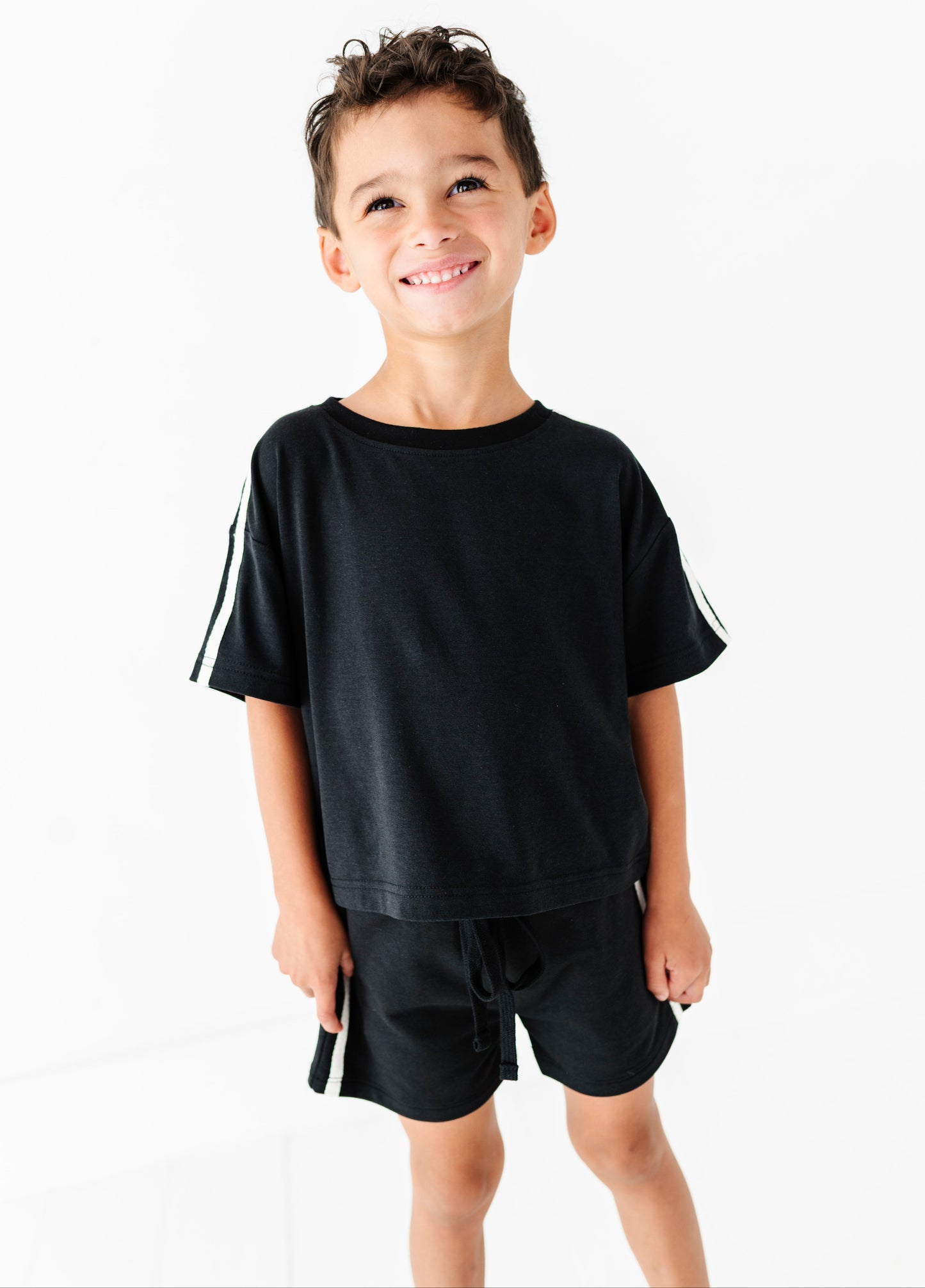 A young boy smiling and wearing a black short-sleeve t-shirt with white stripes on the sleeves and matching black shorts with white side stripes, photographed against a clean white background.