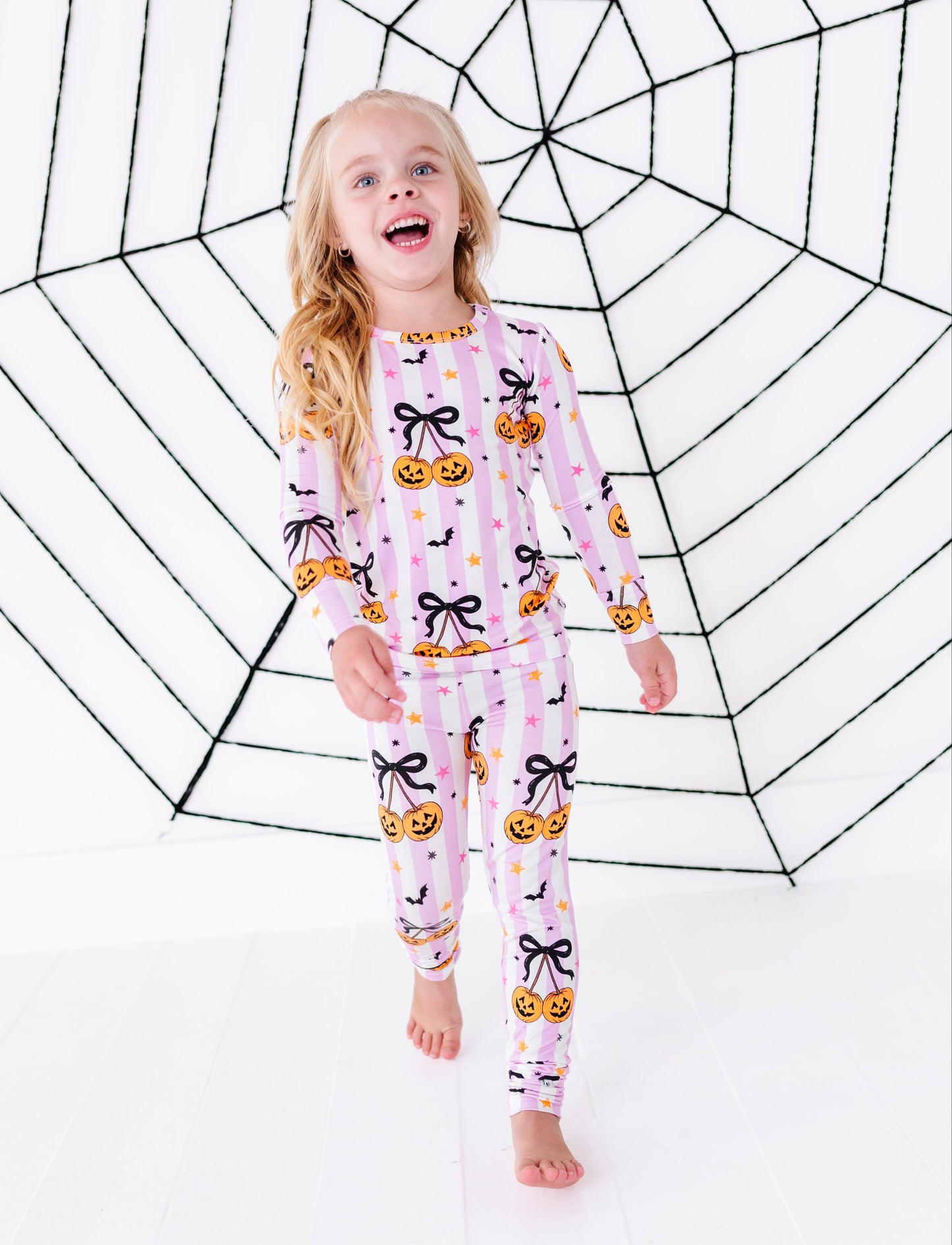 A young girl with blonde hair stands barefoot in front of a large black spiderweb backdrop, wearing a light pink long-sleeve pajama set with a Halloween print of black bows, orange pumpkins, and small yellow stars.