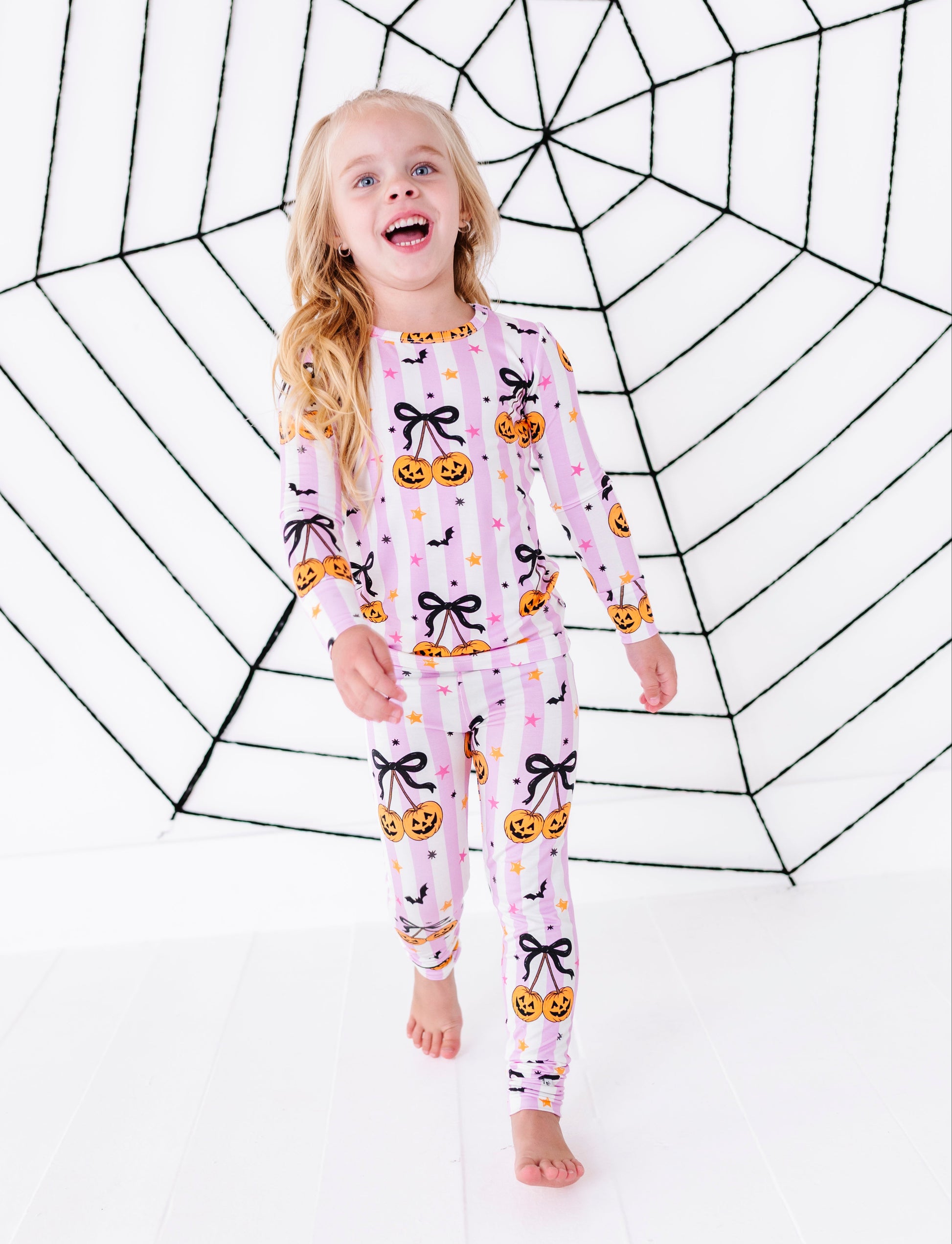 A young girl with blonde hair stands barefoot in front of a large black spiderweb backdrop, wearing a light pink long-sleeve pajama set with a Halloween print of black bows, orange pumpkins, and small yellow stars.