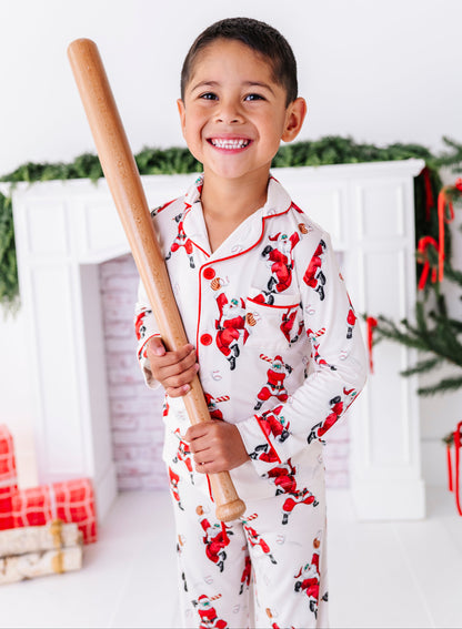 A little boy wearing a loose-fitting bamboo lounge set featuring Santa throwing baseballs and knocking them out of the park! He's holding a wooden bat while standing in front of a fireplace and a blurry Christmas tree