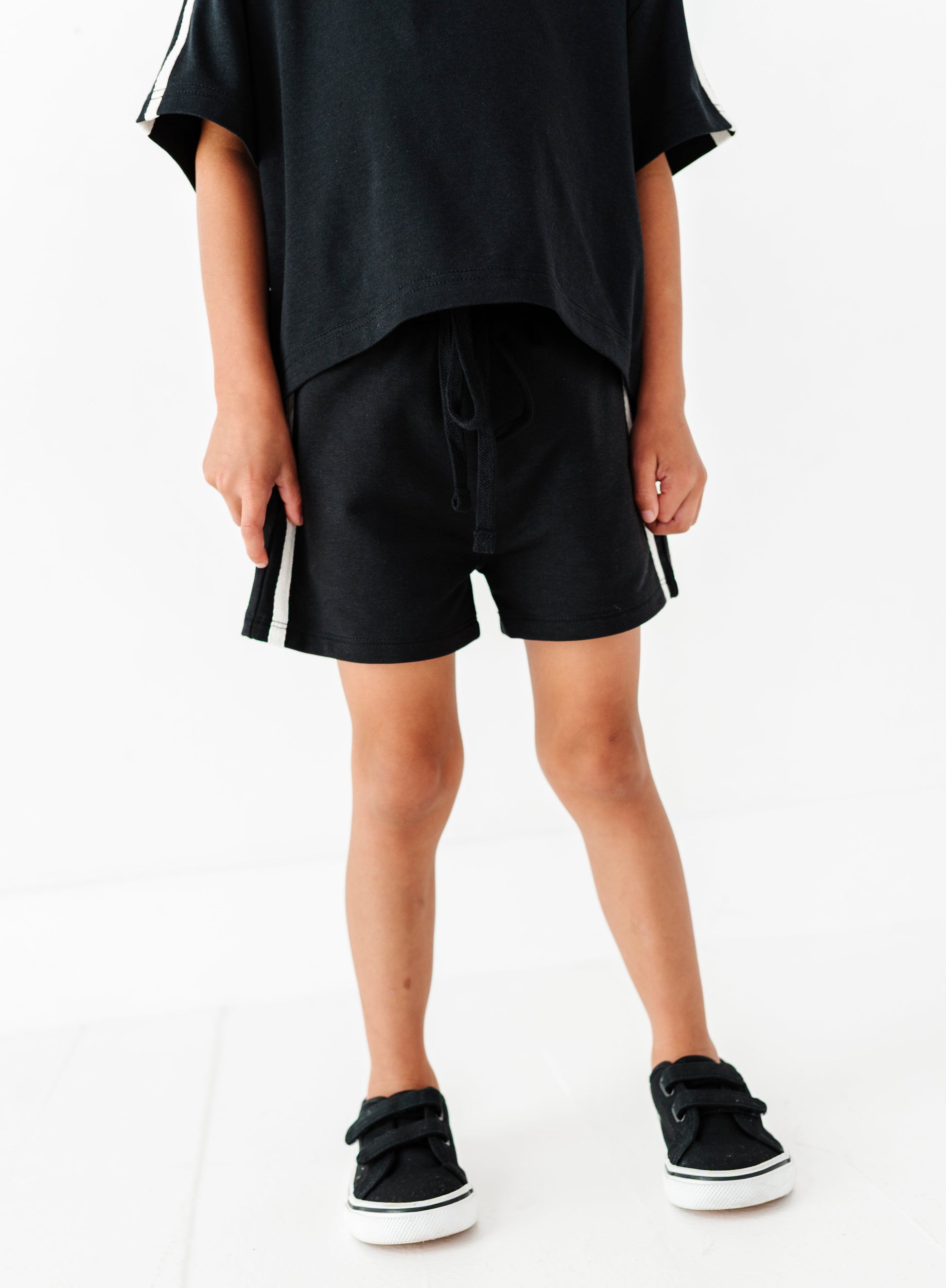 A child wearing black athletic shorts with white side stripes, a black t-shirt, and black sneakers with white soles, standing against a white studio background.