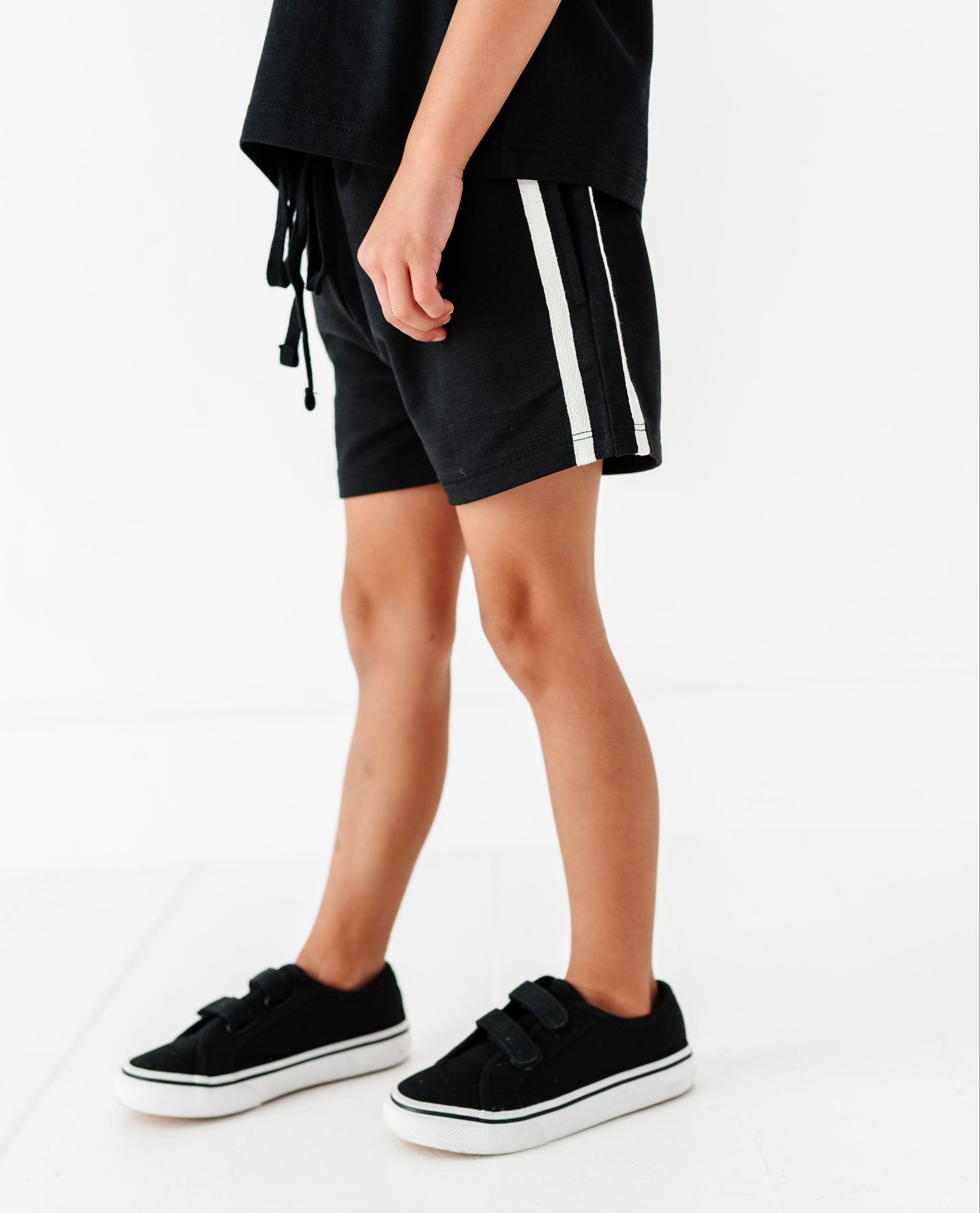 A person wearing black shorts with white side stripes, a loose black top, and black platform sneakers, photographed against a clean white studio background.