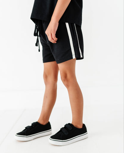 A person wearing black shorts with white side stripes, a loose black top, and black platform sneakers, photographed against a clean white studio background.