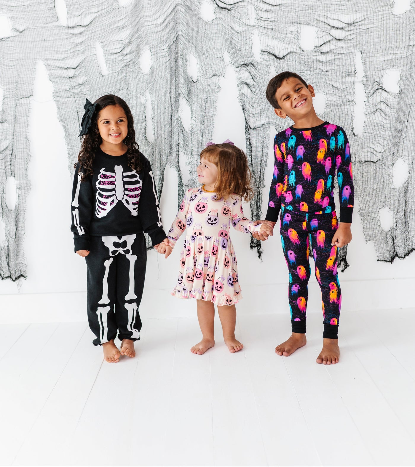 Three children wearing colorful pajamas standing in front of a decorative white wall with black patterns.