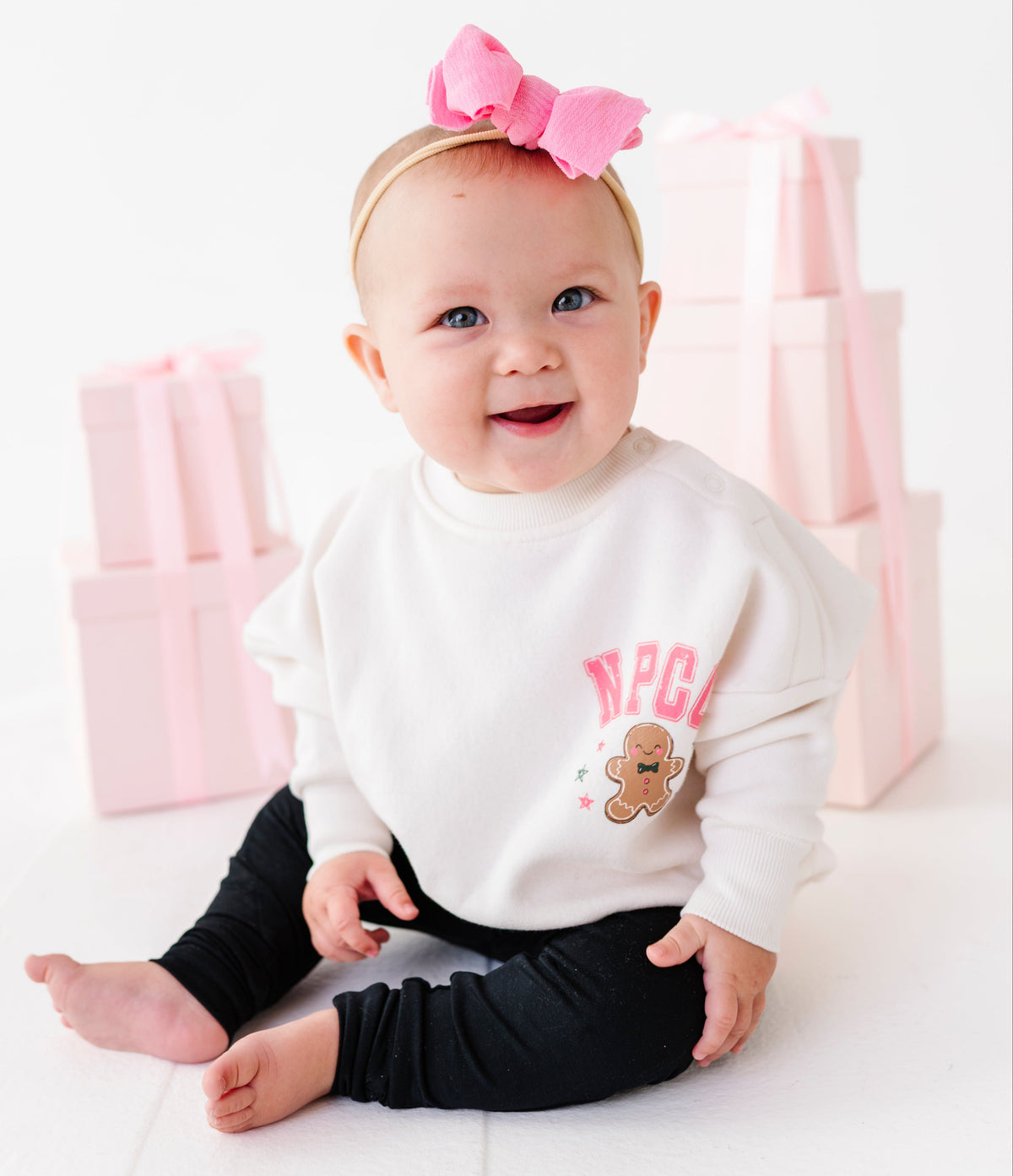 A baby sitting on a white floor wearing a long-sleeve crewneck sweatshirt with pink lettering and a gingerbread cookie graphic, black leggings, and a pink bow headband. Pink gift boxes are stacked in the background.