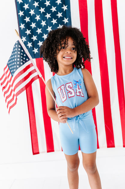 Child wearing a light blue 'USA' tank top and coordinating bamboo biker shorts in front of an American flag