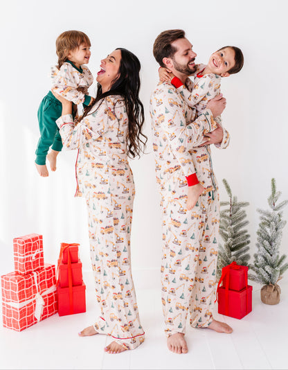 A family of four wearing matching Christmas-themed bamboo pajamas stands together in a bright studio setting. The pajamas have an off-white base with a repeating print of holiday vehicles and trees.