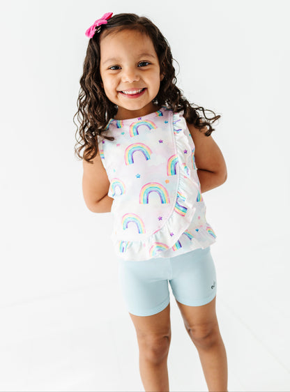 Young girl wearing a sleeveless bamboo peplum top with watercolor rainbow patterns and light blue bamboo biker shorts on a white background