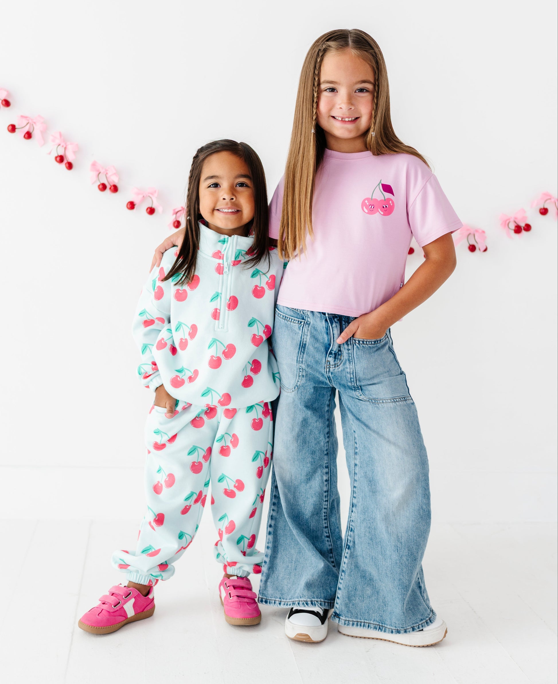 Two young girls modeling matching cherry-themed apparel against a white studio background with a pink cherry garland. The girl on the left wears a light blue and pink cherry print zip-up hoodie and pants set with pink sneakers.