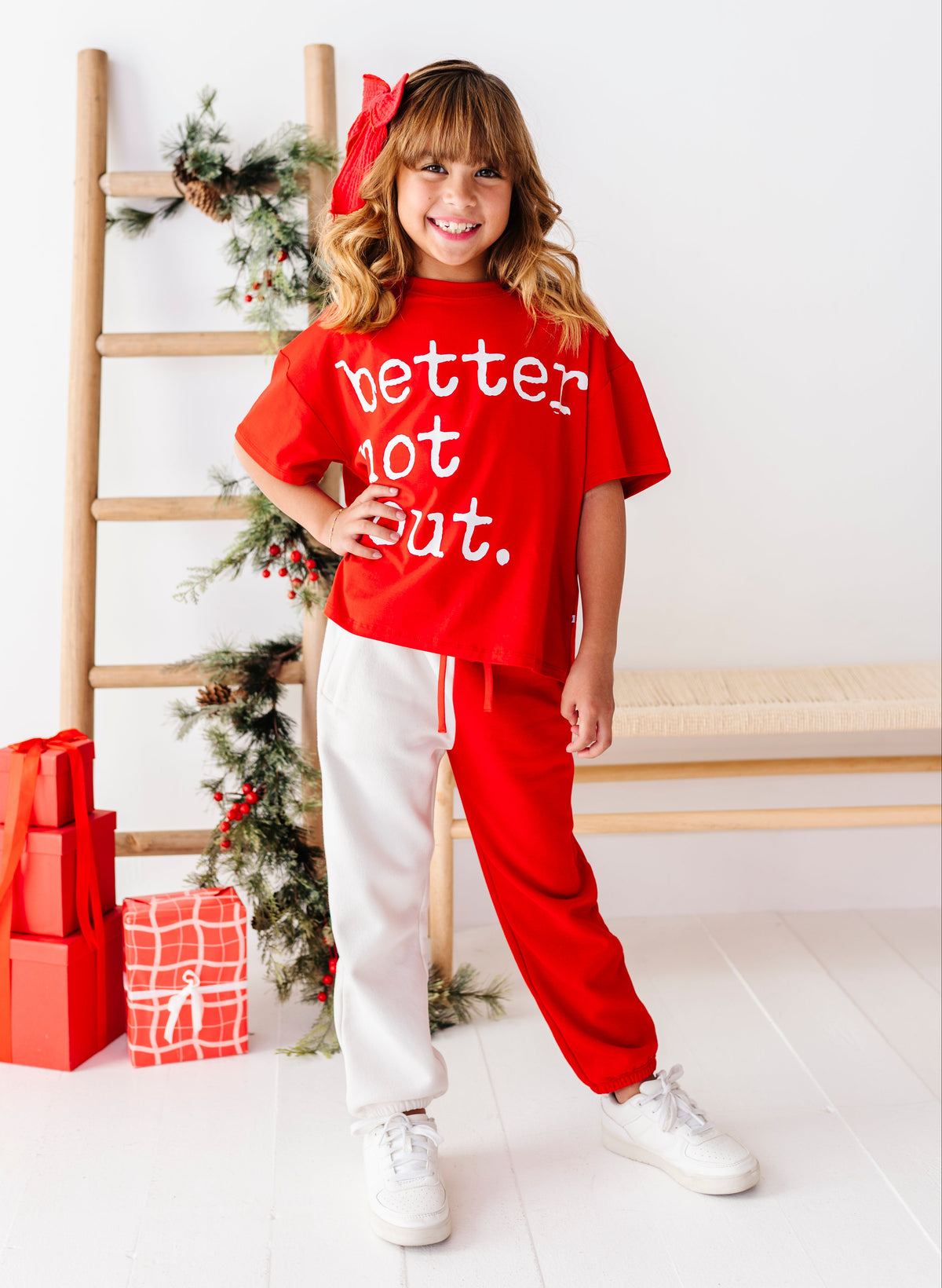 A young girl stands in a bright room wearing a red graphic t-shirt that says 'better lot out.' in white text, paired with white sweatpants and white sneakers.