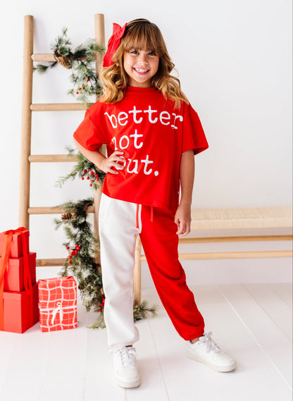 A young girl stands in a bright room wearing a red graphic t-shirt that says 'better lot out.' in white text, paired with white sweatpants and white sneakers.