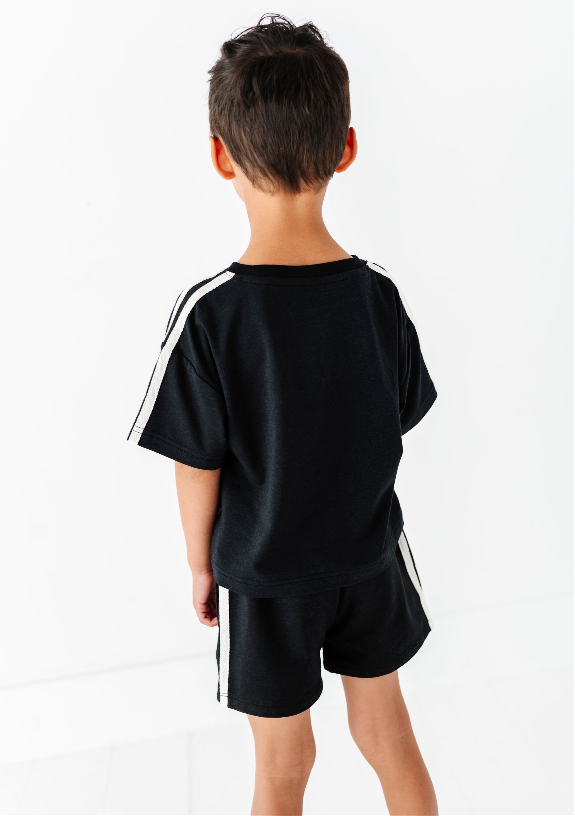 A child viewed from behind wearing a black short-sleeve t-shirt and matching black shorts, both with white side stripes, against a clean white studio background.