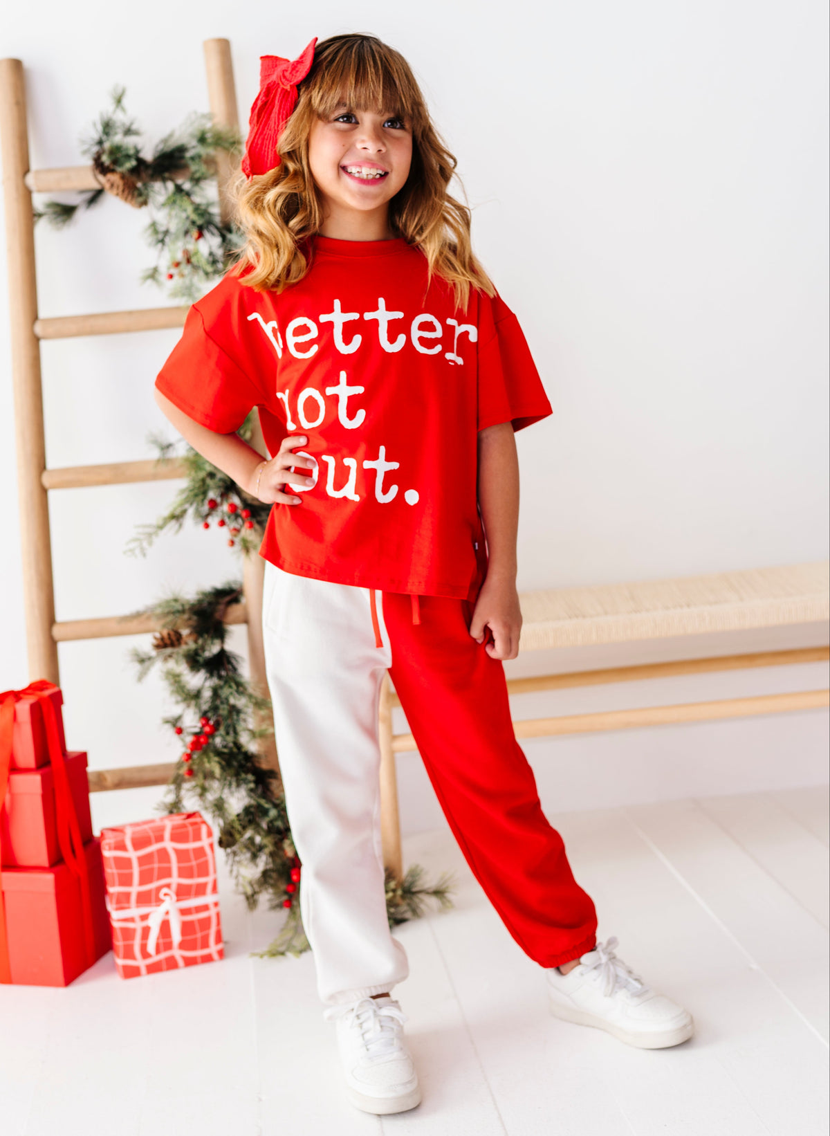 A young girl stands in a bright room wearing a red graphic T-shirt that says 'Better Not Out.' and matching red sweatpants with white drawstrings, paired with white sneakers.