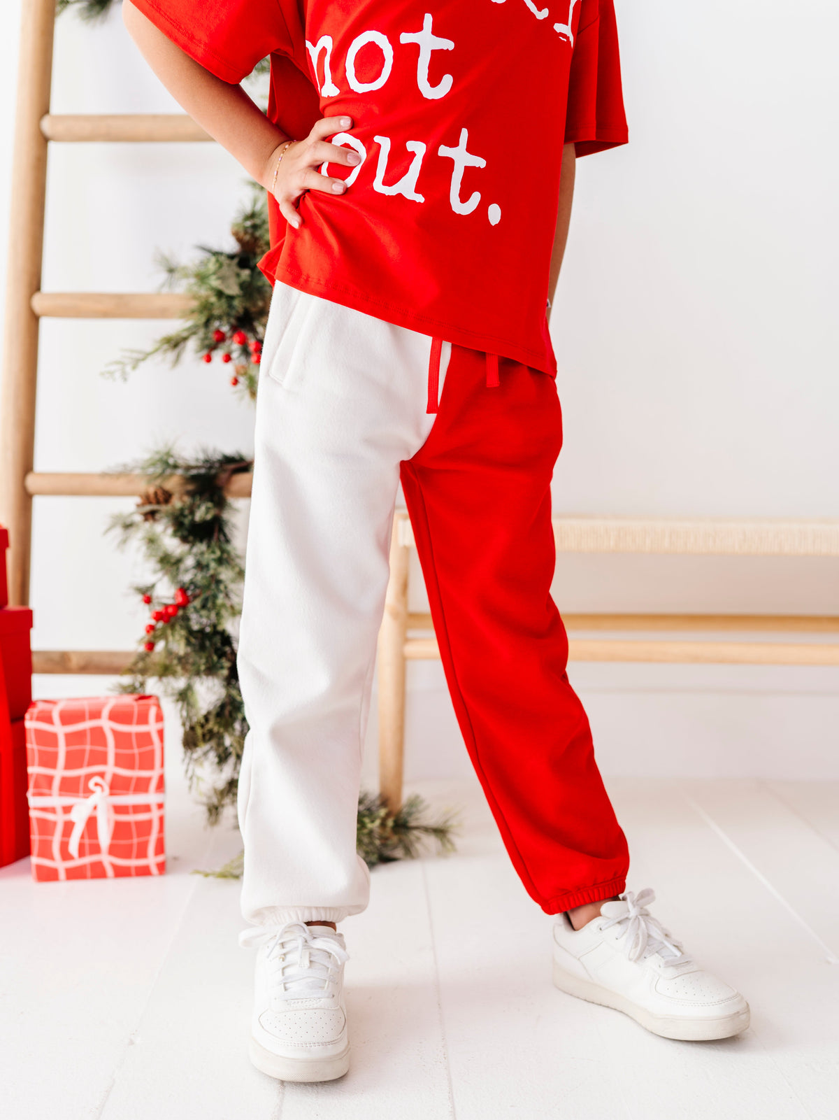 A child wearing a red graphic t-shirt and contrasting red and white sweatpants, standing in a bright room with a wooden ladder and Christmas decorations in the background.
