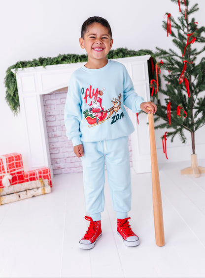 A little boy standing in front of a fireplace holding a wooden baseball bat and wearing a baseball crewneck. The sweatshirt says "no fly zone" and shows Santa catching a baseball in his leather glove 