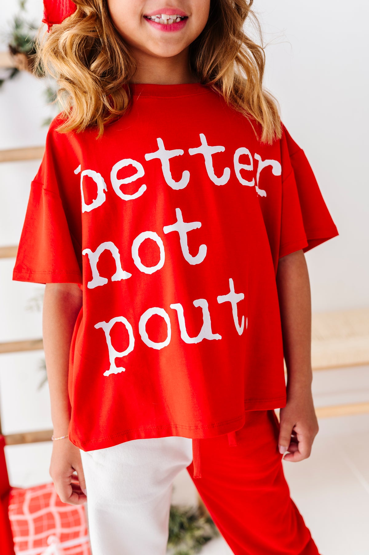 A young girl wearing a red oversized t-shirt with the white text 'better not pout' and matching red pants, shown in a bright indoor setting.
