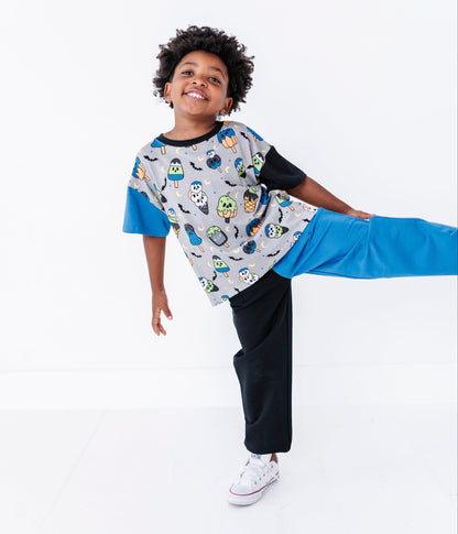 A young boy wearing a short-sleeve t-shirt with a colorful cartoon character print on a gray body and bright blue sleeves, paired with black pants and white sneakers, posing dynamically against a solid white studio background.