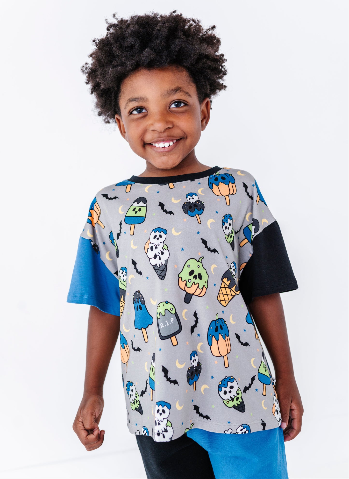 A smiling young boy with curly black hair wearing a gray short-sleeve t-shirt with a colorful Halloween print of popsicles, bats, and skulls, paired with blue and black pants, against a plain white background.
