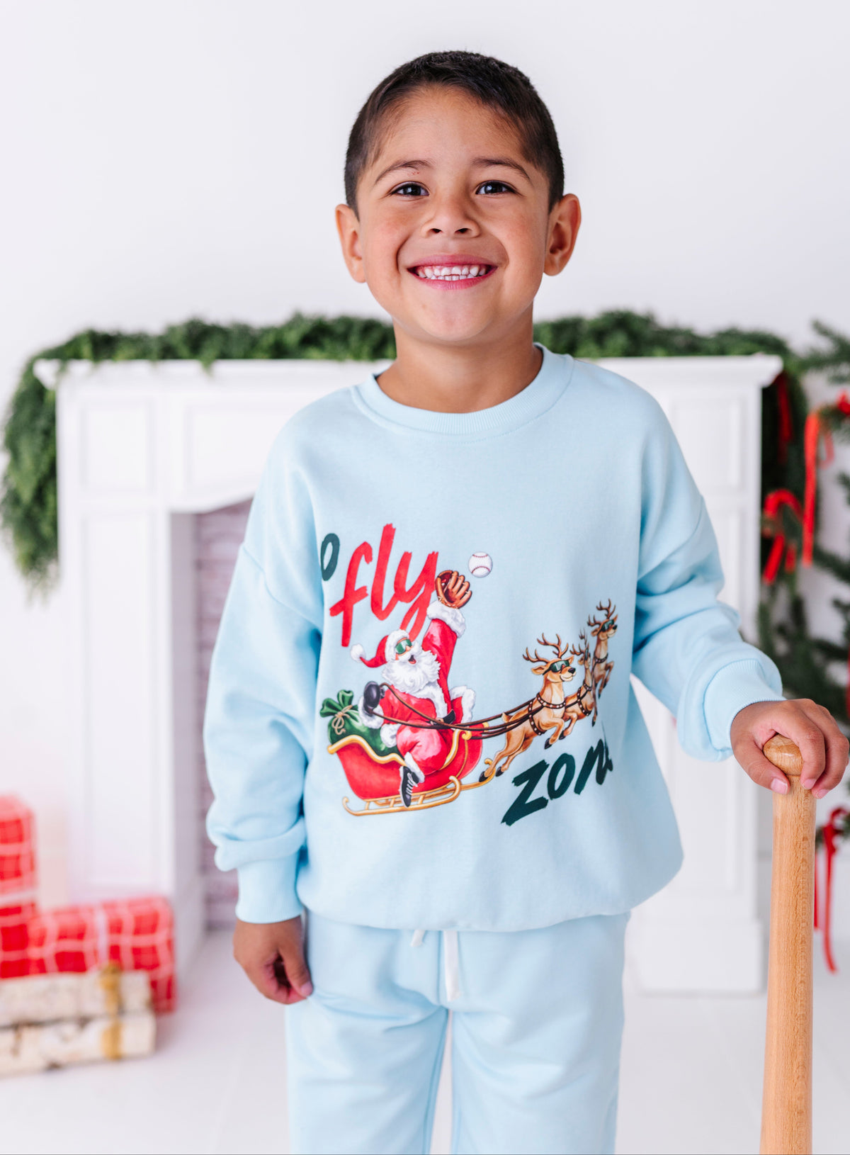 A young boy wearing a blue baseball crewneck that says "no fly zone" with Santa in a sleigh catching a baseball. He's holding a wooden bat and standing in front of a fireplace 