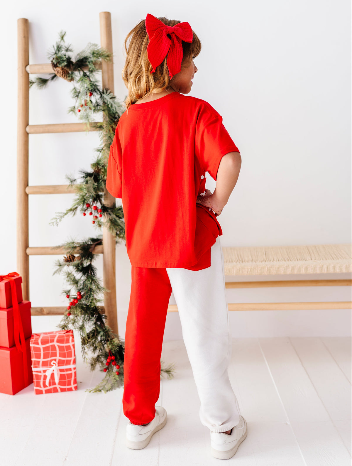 A child stands with their back to the camera wearing a festive holiday outfit consisting of a bright red short-sleeve top, matching red pants, and a large red bow headband. The background is a bright white room decorated with a wooden ladder, green pine garland, and wrapped presents.