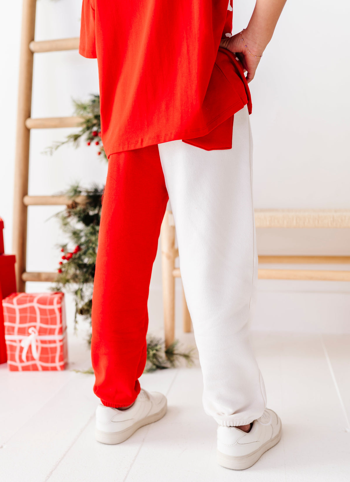A person shown from the back wearing a bright red oversized top and a split-color outfit with one leg in red and the other in white, paired with white sneakers. The background is a bright, minimalist room with a wooden ladder and a small decorated Christmas tree.