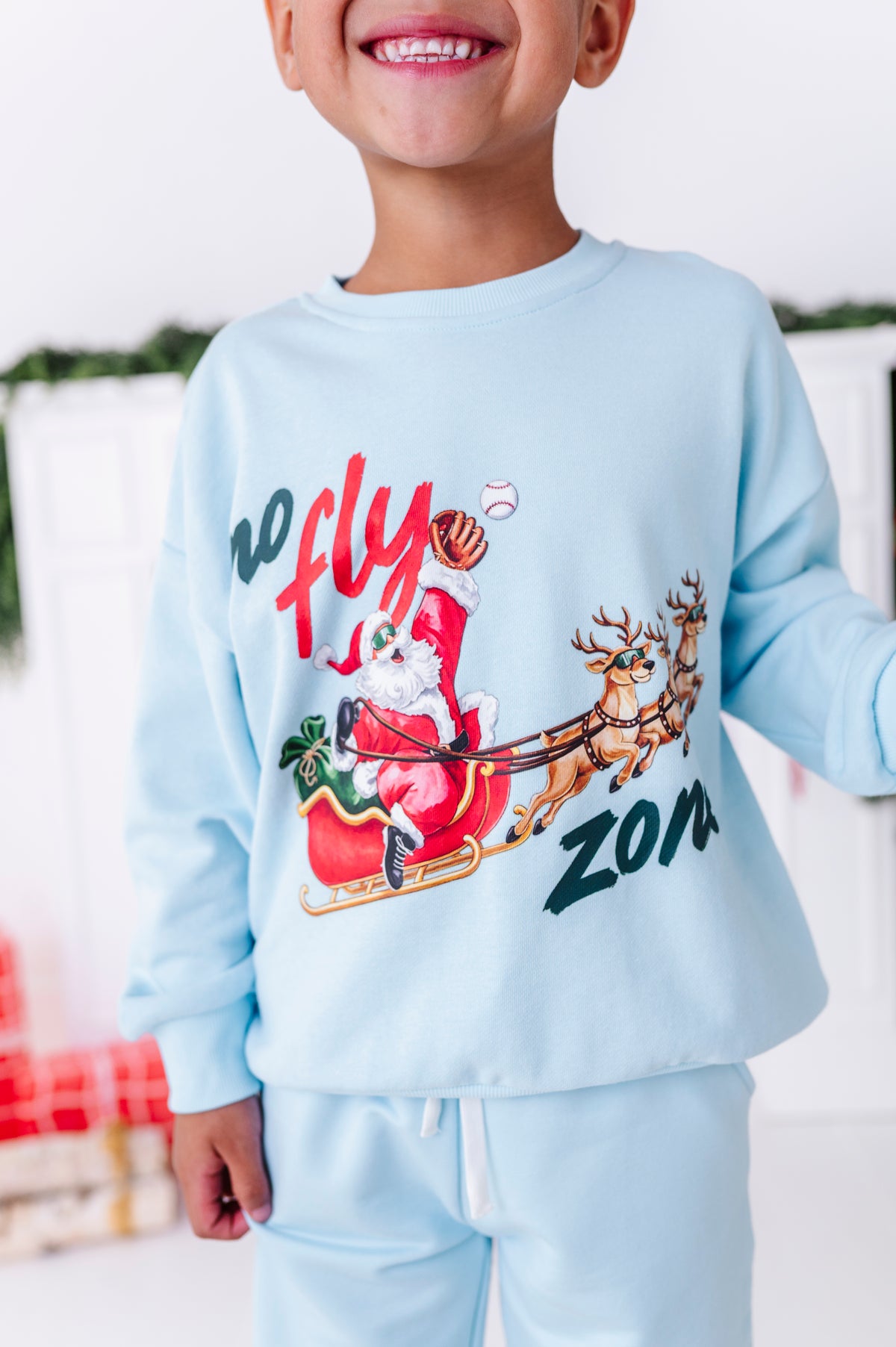 A close-up shot of the light blue baseball crewneck featuring "no fly zone" with Santa catching a baseball while flying in his sleigh 