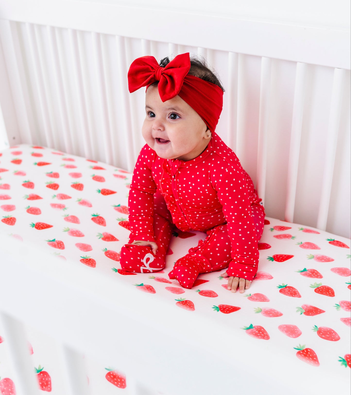 Baby in red outfit with large bow sitting on a crib with strawberry patterned sheet.