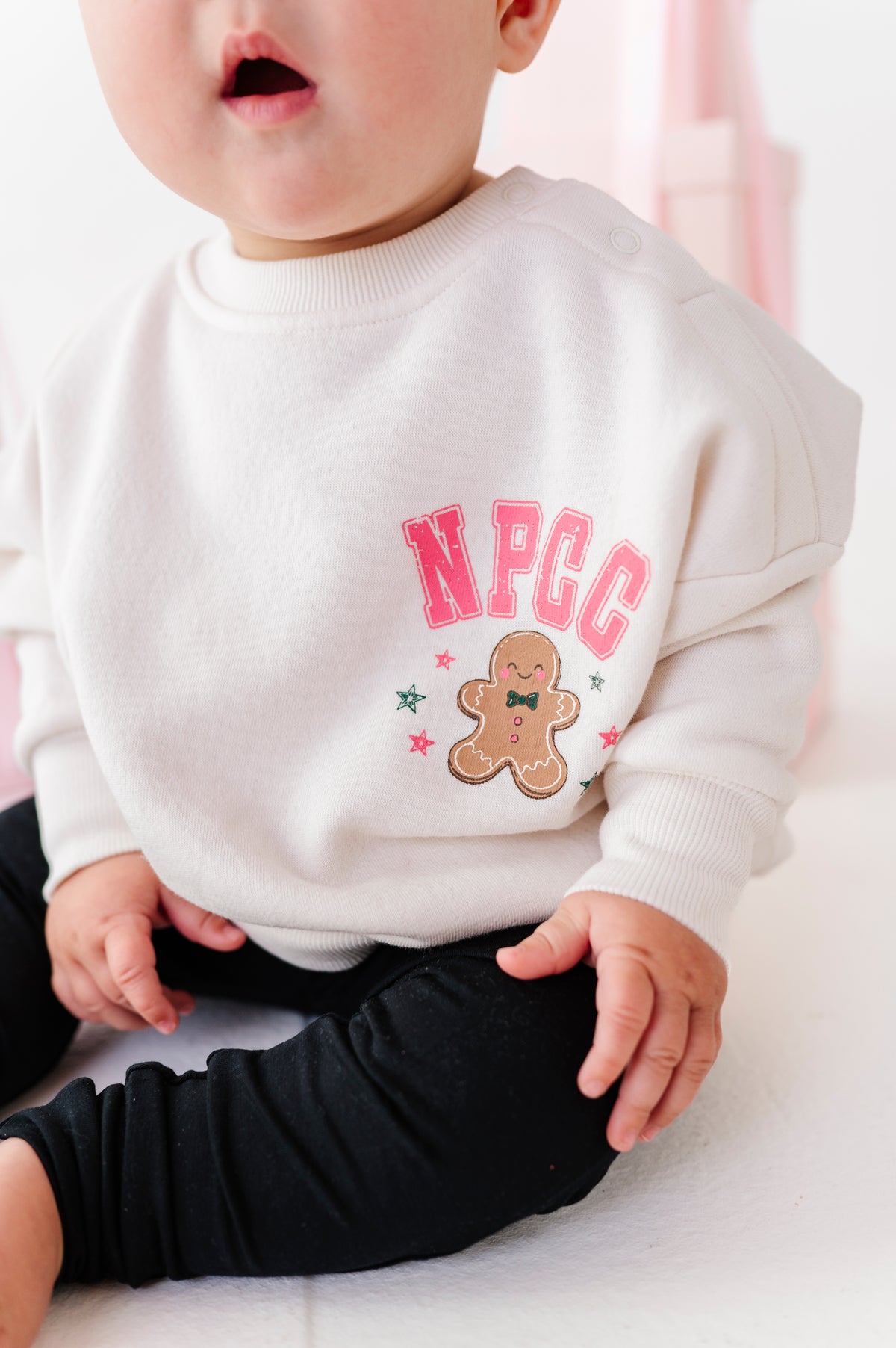 A baby wearing a cream-colored long-sleeve crewneck with a pink 'NPCC' graphic and a gingerbread man design, paired with bamboo leggings.