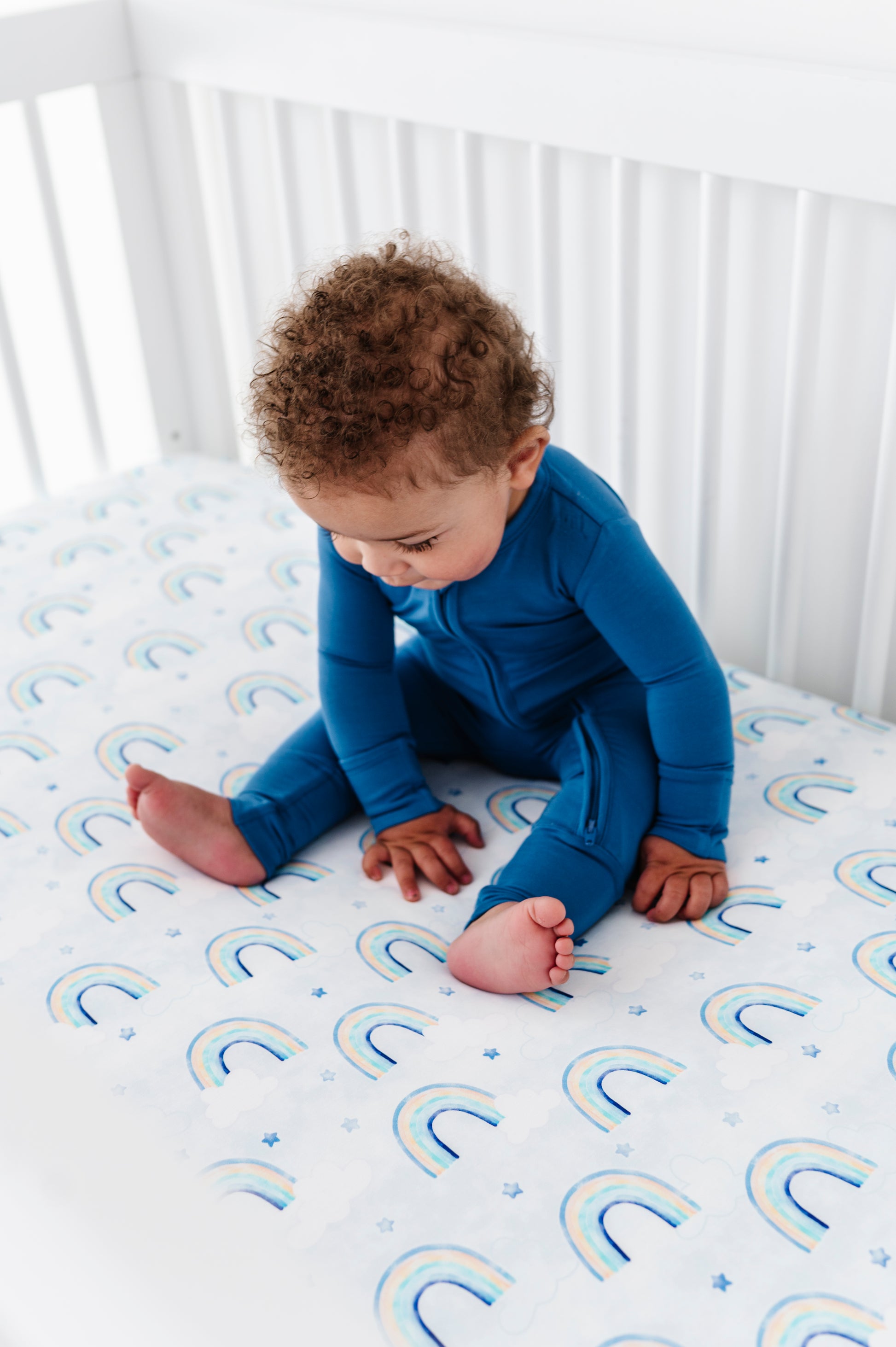 Baby in a blue bamboo zip romper sitting on a crib mattress with rainbow patterns.