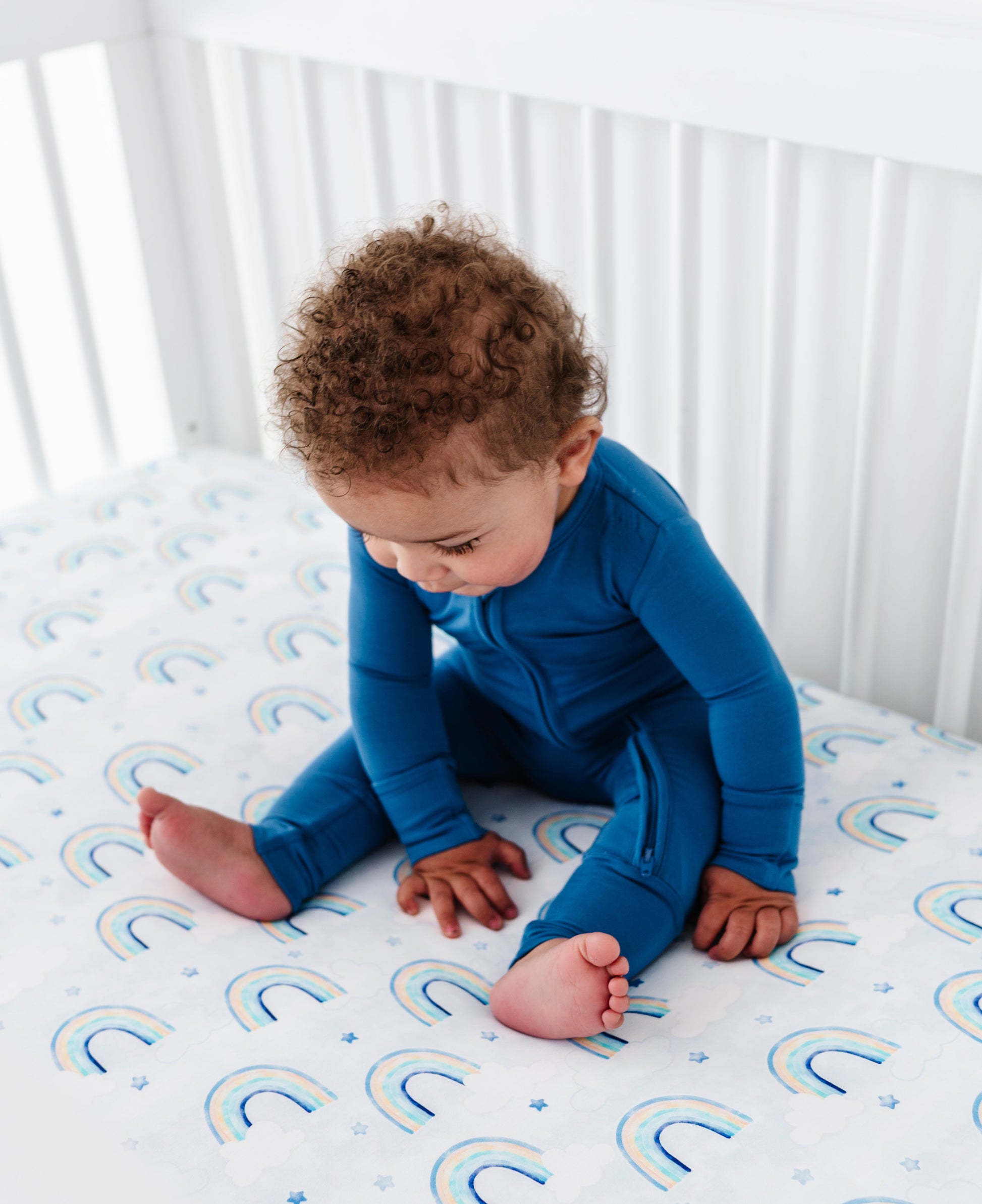 Baby in a blue convertible romper sitting on a crib mattress with rainbow patterns.