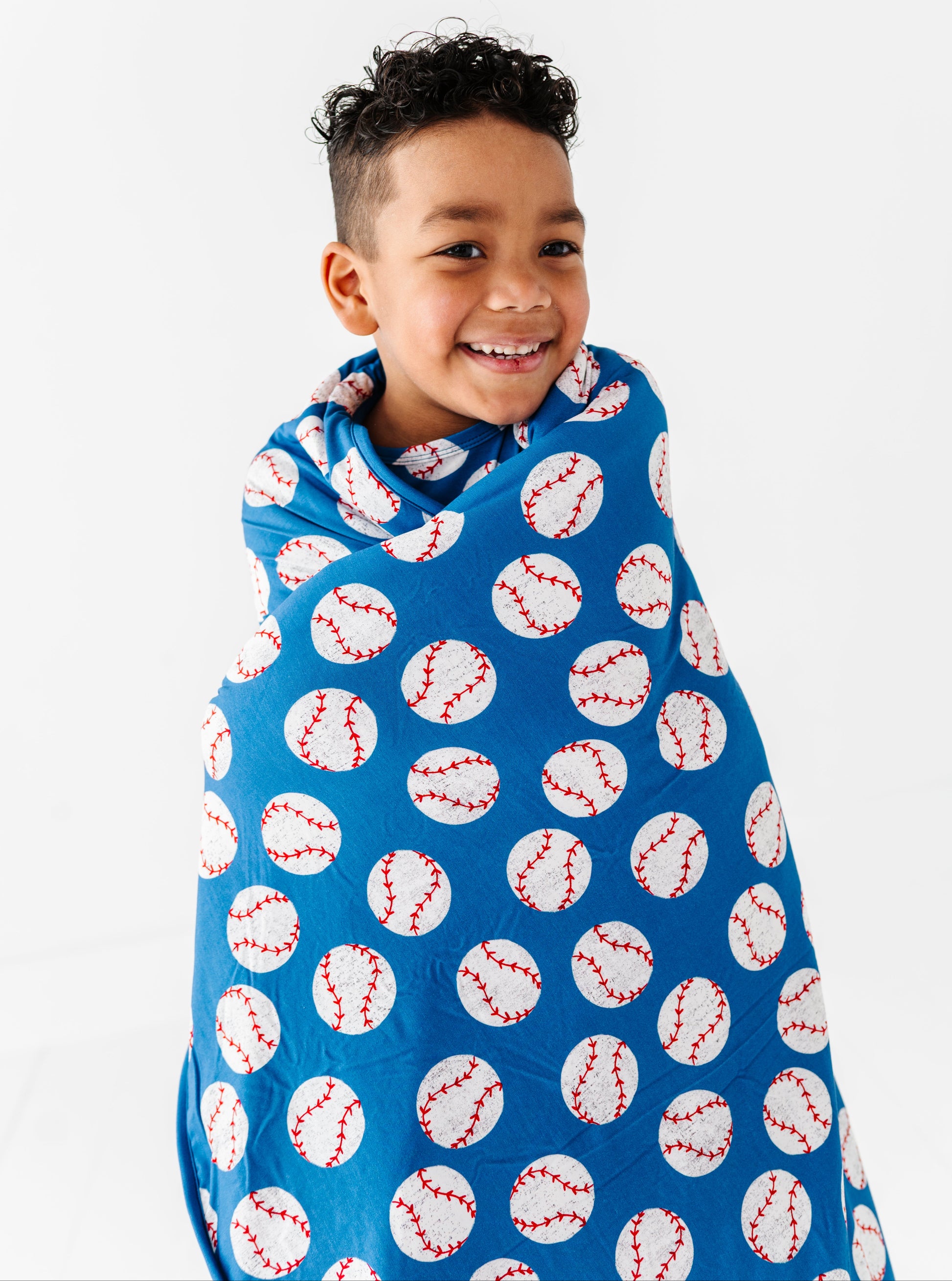 Little boy wrapped in a blue baseball bamboo blanket against a white background
