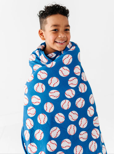 Little boy wrapped in a blue baseball bamboo blanket against a white background