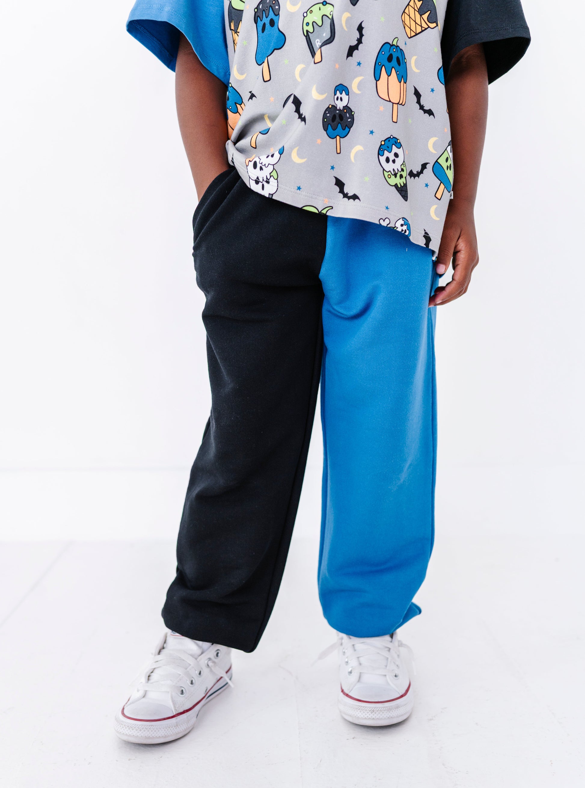 A child wearing half black and half bright blue sweatpants, a gray patterned t-shirt with a blue sleeve, and white sneakers, shown from the waist down against a white studio background.