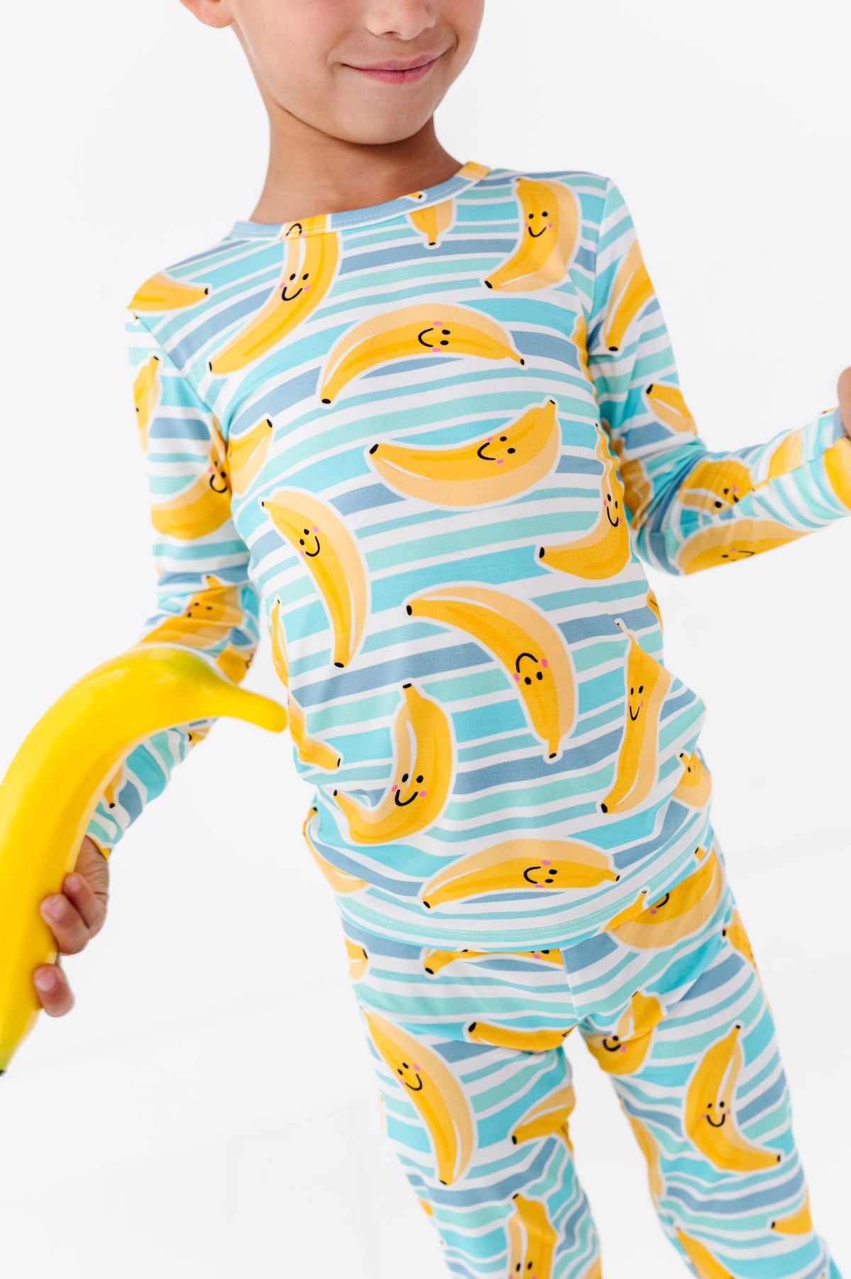 A child wearing a long-sleeve pajama set with a playful banana print, holding a yellow banana prop against a clean white background.