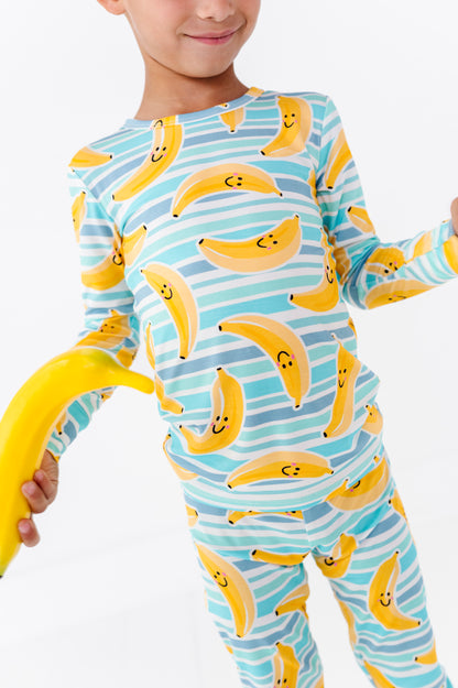 A child wearing a long-sleeve pajama set with a playful banana print, holding a yellow banana prop against a clean white background.