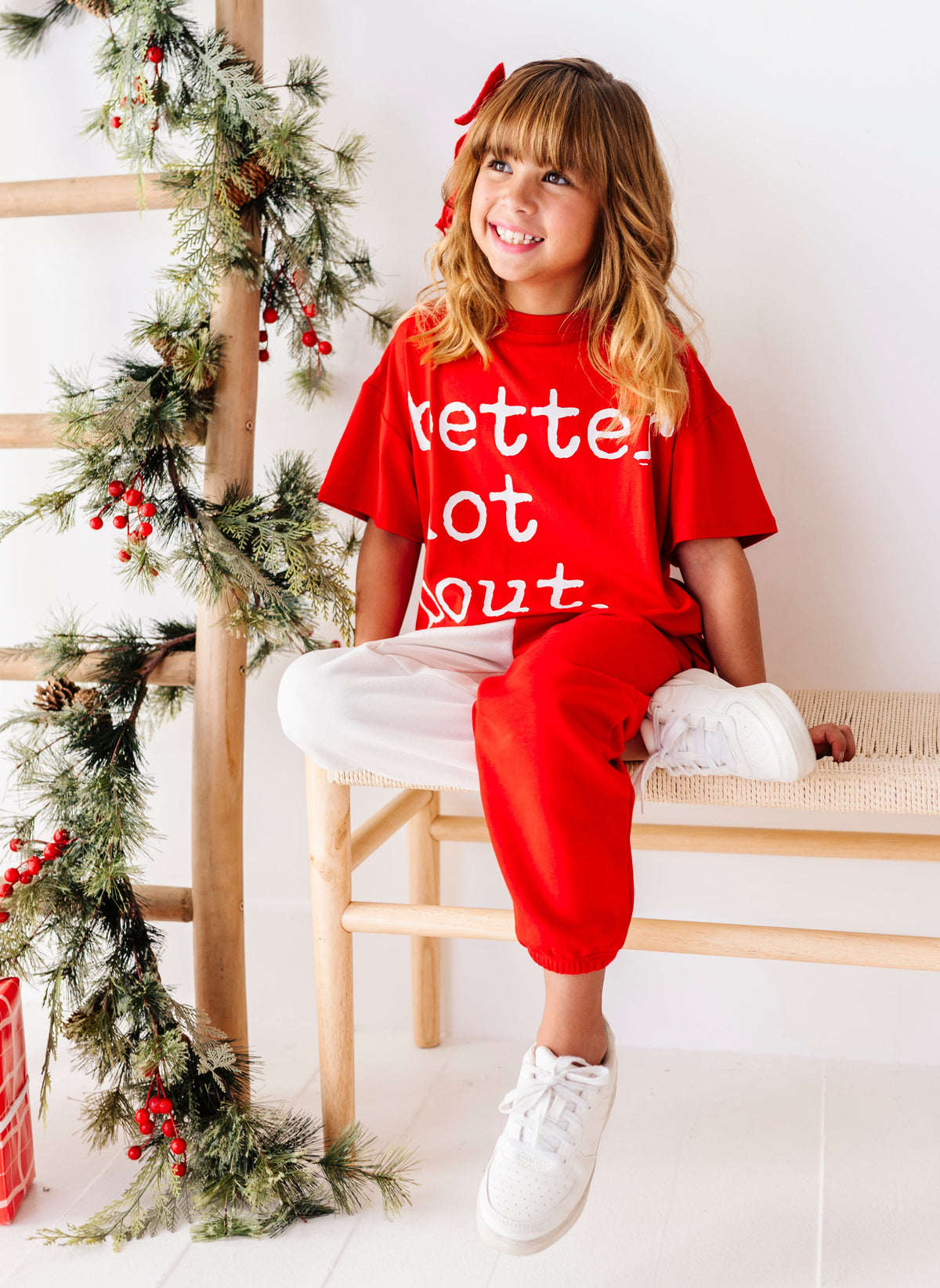 A young girl with blonde hair and a red bow sits on a wooden bench wearing a red Christmas-themed t-shirt with white text that reads 'kette out' and matching red sweatpants. The background is a bright white wall with a wooden ladder decorated with a green pine garland and red berries.