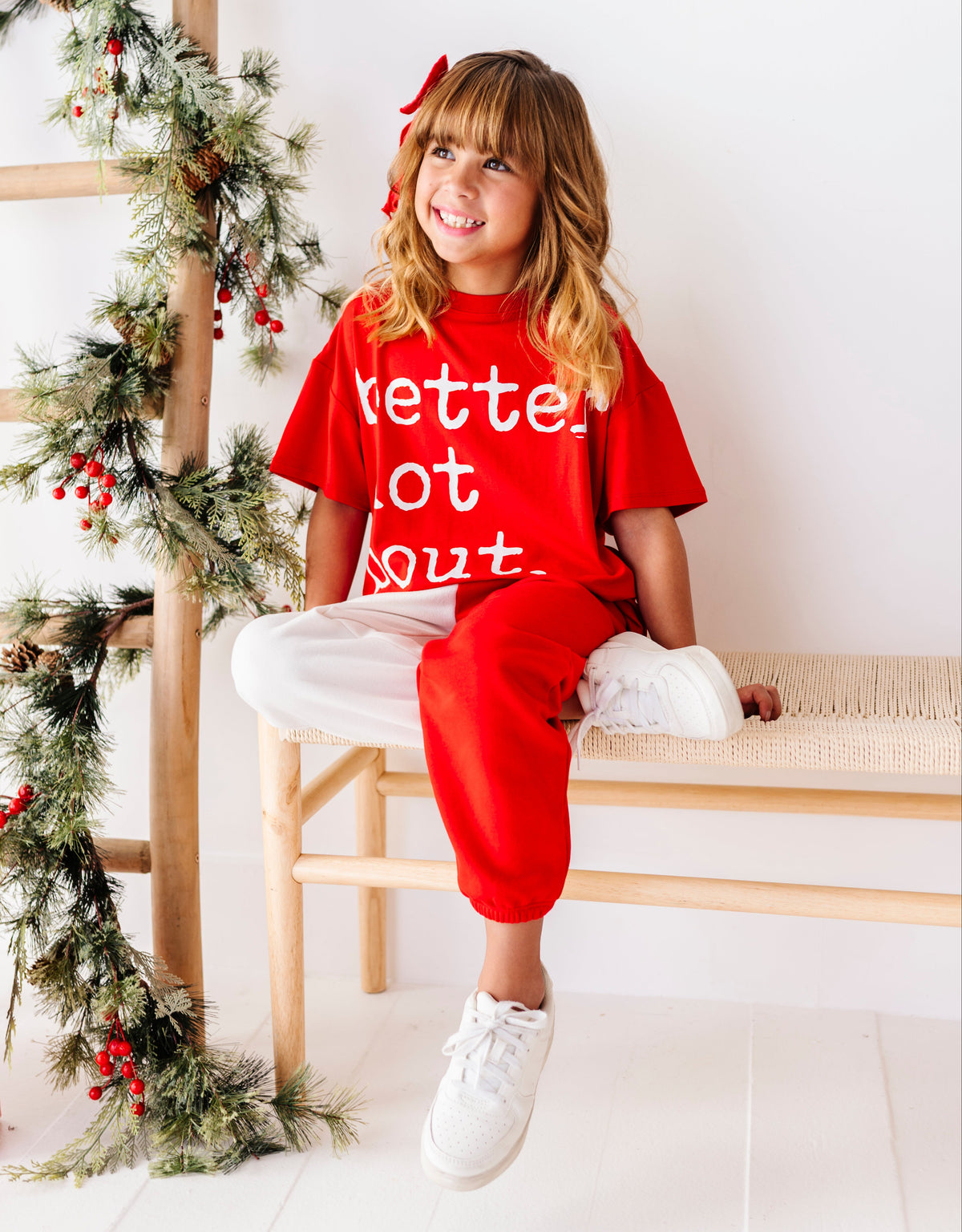 A young girl with blonde hair and a red bow sits on a wooden bench wearing a red Christmas-themed t-shirt with white text that reads 'kette out' and matching red sweatpants. The background is a bright white wall with a wooden ladder decorated with a green pine garland and red berries.