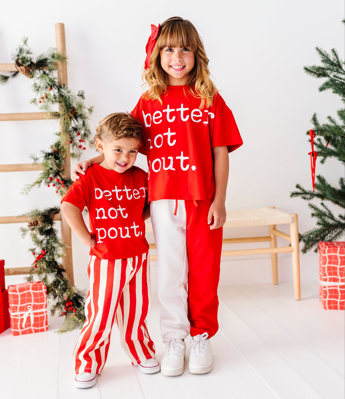Two children standing together in a bright white room decorated for Christmas, wearing matching red holiday sweatshirts with white text that reads 'better not out.' and red-and-white striped pants.
