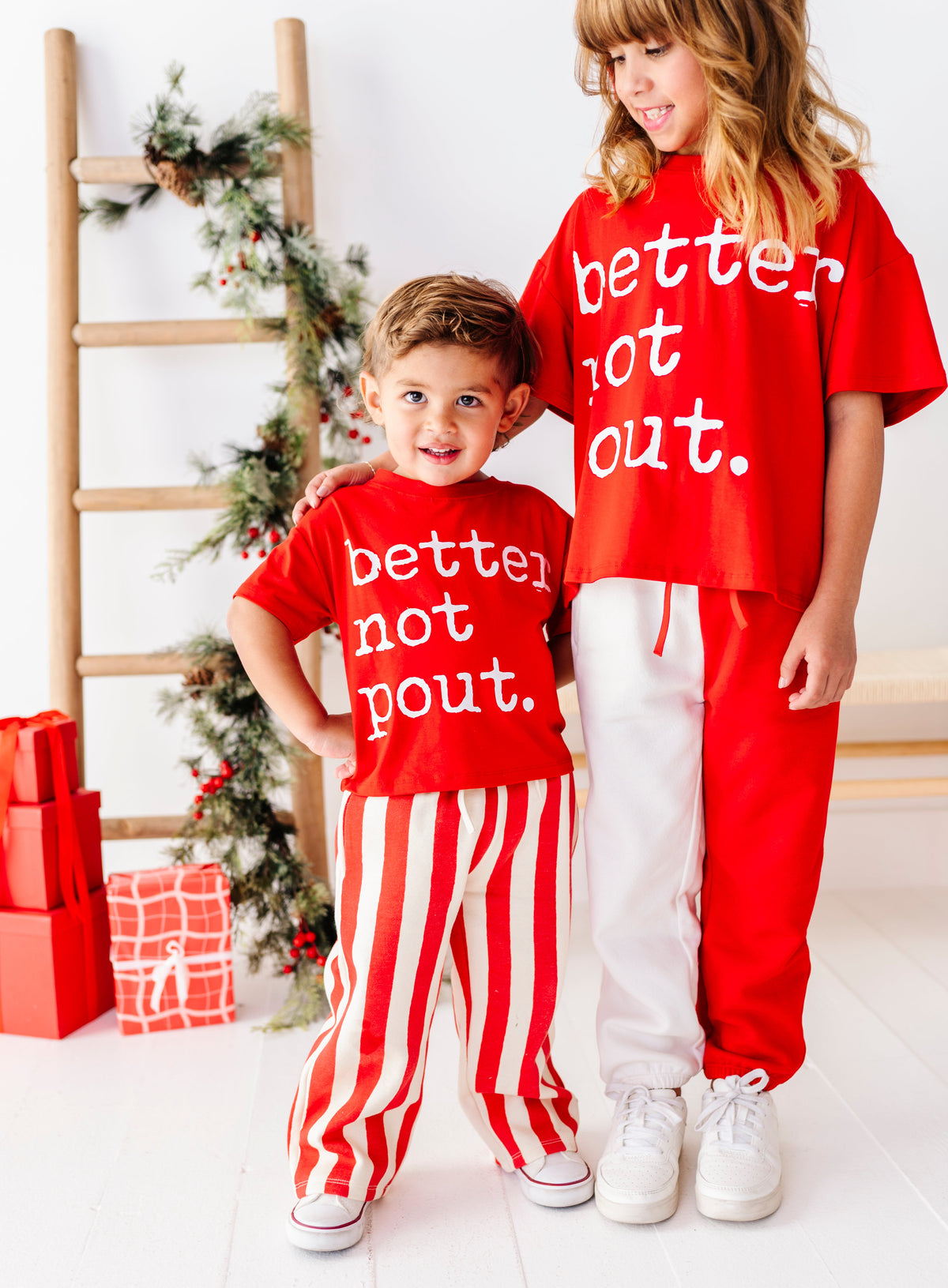 A young boy and a girl wearing matching red Christmas-themed sweatshirt and pants sets with the text 'better not out.' in white, standing in a bright room with a wooden ladder and holiday decorations.