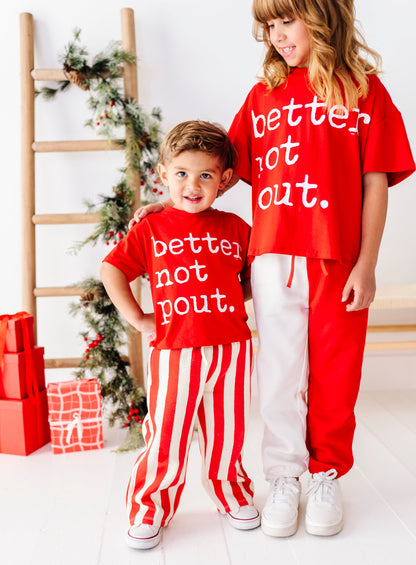 A young boy and a girl wearing matching red Christmas-themed sweatshirt and pants sets with the text 'better not out.' in white, standing in a bright room with a wooden ladder and holiday decorations.