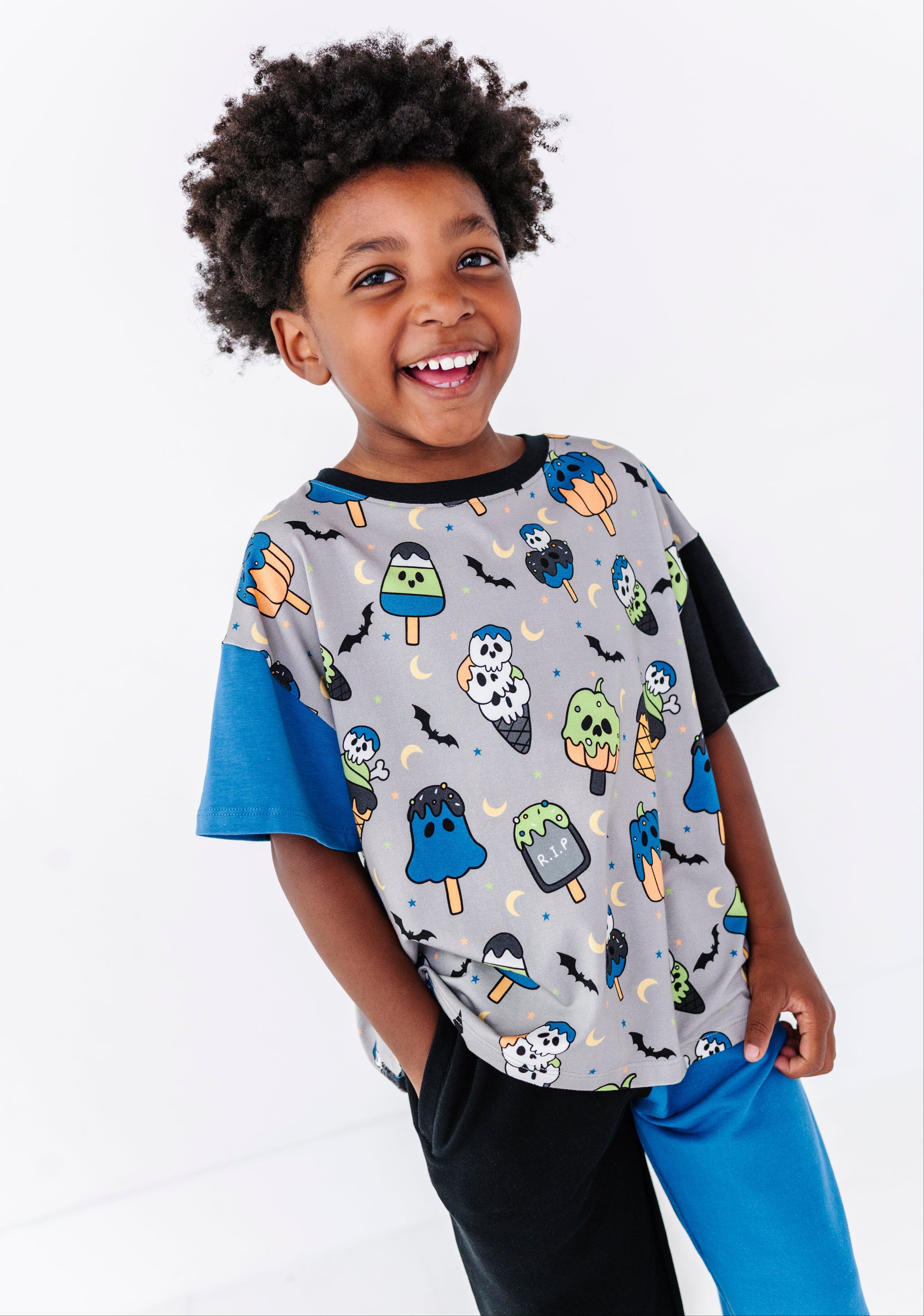 A young boy with curly dark hair smiling and looking to the side, wearing a short-sleeve t-shirt with a gray body covered in a colorful Halloween popsicle print, blue sleeves, and black pants, against a plain white background.