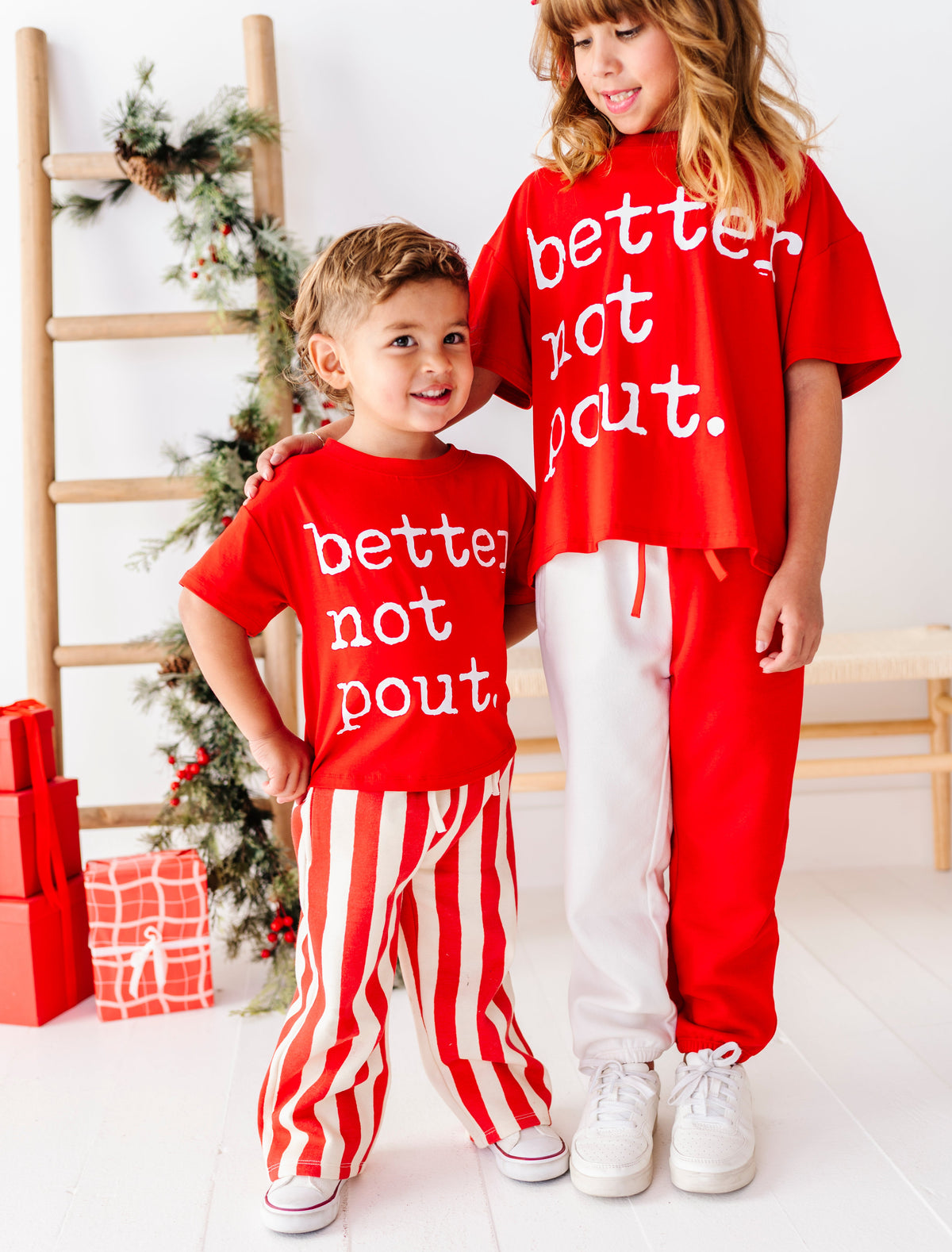 Two children modeling matching red holiday outfits with white text reading 'better not pout.' in a bright indoor Christmas setting with a ladder, wrapped gifts, and a decorated tree.