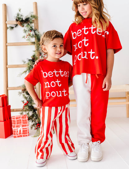 Two children modeling matching red holiday outfits with white text reading 'better not pout.' in a bright indoor Christmas setting with a ladder, wrapped gifts, and a decorated tree.
