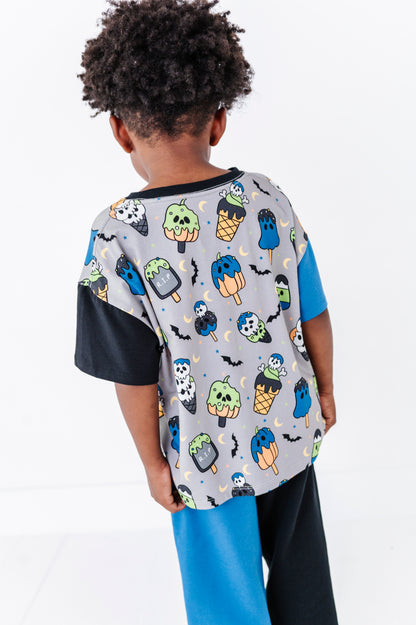A child shown from the back wearing a short-sleeve colorblock t-shirt with a light gray body, black sleeves, and a colorful Halloween-themed ice cream cone print, paired with bright blue and black pants.