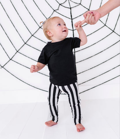 A toddler standing on a white floor in front of a large black spiderweb backdrop, wearing a black short-sleeve top and black-and-white vertically striped pants, reaching up to hold an adult hand.